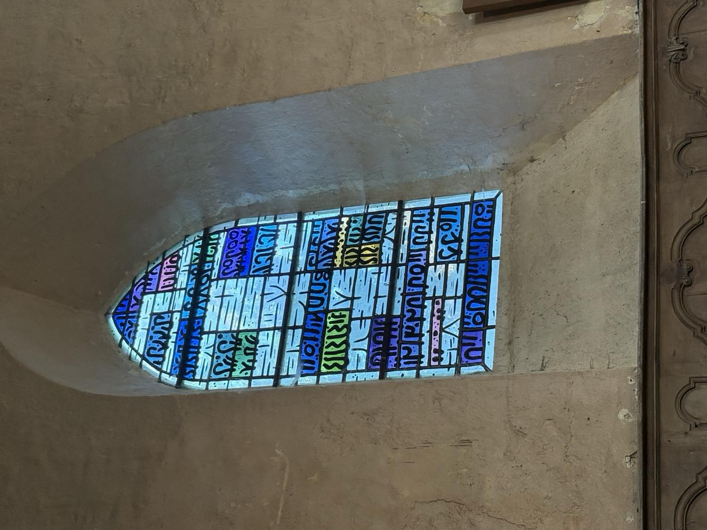 Vitrail en ogive aux verres bleus, violets et verts, éclairant le mur de pierre de la nef.